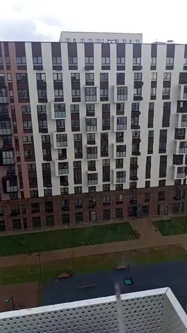 This media file shows a high-rise residential building in Misaylovo, Russia. The building has a modern design with a façade featuring alternating vertical bands of white and brown, with numerous windows and balconies. The lower floors appear to be clad in brick. In front of the building, there is a courtyard area with green spaces, walkways, benches, and a children's playground. In the background, another building is visible, also appearing to be a residential structure. The sky is cloudy with patches of blue, suggesting it could be daytime with variable weather. There are no people visible in the frame. The overall impression is of a residential complex in an urban or suburban setting. No visible text is present.