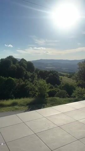 The video shows a panoramic view of a landscape under a bright sun and a blue sky with scattered clouds. In the foreground is a tiled patio area, from which the camera looks out over lush green trees and rolling hills. Distant mountains are visible on the horizon. Power lines stretch across the sky. As the video pans slightly, a road and a blue park bench appear on the right side of the frame. The overall scene evokes a sense of calm and natural beauty.