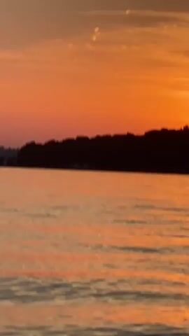 The video shows a body of water with a sunset in the background. The sky is orange and there are clouds. The sun is setting behind a silhouetted landmass. There are ripples on the water. The camera is moving slightly, giving the impression of being on a boat.