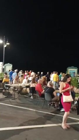 A wide shot captures an outdoor gathering at night, possibly a restaurant or event. The scene is illuminated by artificial lights, including streetlamps and possibly lights from the venue. Numerous people are seated at tables, engaged in conversation or dining. Several individuals are standing and walking around. In the foreground, two women are walking away from the camera. One woman is wearing a red dress and carrying a white purse. The other woman is wearing a white top and light-colored pants, with a red bag slung over her shoulder. Behind them, a man in a black shirt is walking towards the camera, with his back to the viewer. The background is dark, indicating it is nighttime. The ground appears to be asphalt, with white lines painted on it, suggesting a parking lot or pathway. Lush green foliage is visible in some areas, adding to the outdoor ambiance.