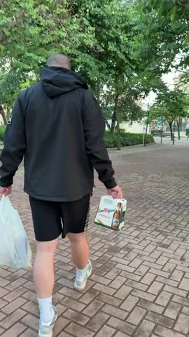 A man walks away from the camera along a brick pathway, holding a six-pack of San Miguel Especial beer in one hand and a white plastic bag in the other. He is wearing a black hooded jacket, black shorts, and gray sneakers with white socks. The setting appears to be a park with trees, grass, and benches visible in the background. The path is lined with trees on both sides. The weather seems mild with good lighting.