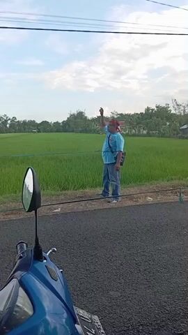A man in a light blue shirt, jeans, and a red headband is walking along the side of a road next to a green field, waving. The road is paved with black asphalt. The camera is mounted on a blue scooter. The field stretches out behind the man, with trees and a few buildings visible in the distance. Overhead are power lines. The sky is blue with scattered white clouds. The man raises his hand several times as he walks.