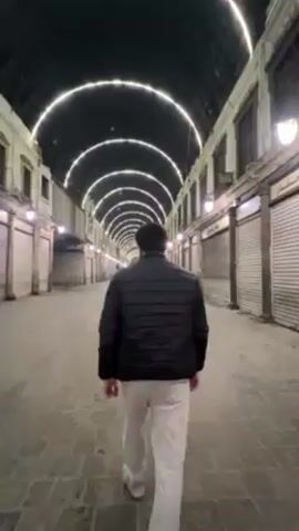 A man walks down a narrow, empty, covered street at night. The street is paved with brick and lined with closed shops on either side. The shops have roll-down metal shutters. Above the street is a series of arched frames illuminated with white lights. Streetlights provide additional illumination. The man is wearing a black jacket, white pants, and white sneakers. He turns his head slightly to the side while walking towards the camera.