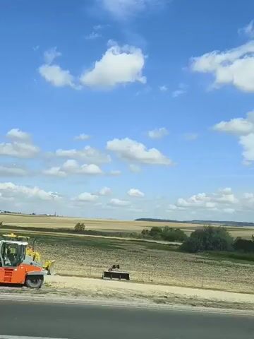 A rural landscape is viewed from a moving vehicle. The sky is blue with scattered white clouds. In the foreground, a gray pickup truck is parked on the side of a road. In the middle ground, a large, dry field stretches out, with some trees and bushes on the right. In the distance, more fields and trees are visible. A second vehicle, a black car, drives by in the middle ground. The video appears to be taken during the daytime.