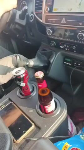 A video shows the interior of a vehicle from the passenger seat. The dashboard is visible, with a GPS screen displaying a map and navigation route. There are two drinks in the cup holders in the center console: a red soda can and a brown glass bottle with a red label. The camera pans to the right, showing the road ahead and the surrounding landscape of trees and fields. The video appears to be taken while the vehicle is in motion.