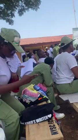 Several individuals dressed in light green uniforms, reminiscent of military or civil service attire, are gathered outdoors, possibly for an official function. They are seated on wooden benches, indicating a temporary or informal setting. One person is holding a child, documents, and a phone. A sign for "National Youth Service Corps" is visible in the background, suggesting a youth service or civic engagement program. Other items such as a bag with "THE LORD'S WRITER" inscribed, and piles of colorful clothes are scattered around, indicating that this is a personal gathering. The overall environment suggests a casual, outdoor event, potentially involving youth or community service activities.