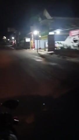 The video shows a first-person perspective of riding a motorcycle at night on a paved road. The rider's hands are visible on the handlebars, and the dashboard of the motorcycle is partially in view. The road is illuminated by streetlights and the headlights of the motorcycle, creating a warm glow. On either side of the road, there are buildings, some with lights on, and trees. The scene appears to be a quiet street in a town or village.