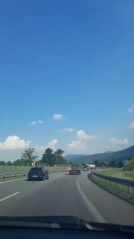 The video captures a scenic drive through a mountainous region on a sunny day. The camera is mounted inside a vehicle, providing a first-person perspective of the journey. The highway ahead is mostly clear, with a few cars visible, including a black sedan in the left lane and a light-colored SUV or truck in the right lane. The road is bordered by lush green trees and vegetation, with rolling hills and mountains in the background. The sky is a vibrant blue, dotted with fluffy white clouds, suggesting fair weather.

A prominent detail is a series of red and white directional signs on the left side of the road, indicating a curve. The presence of guardrails along the roadside further emphasizes the highway setting. The overall atmosphere appears peaceful and relaxed, characteristic of a pleasant road trip through a natural landscape. The time of day is clearly daytime, judging by the bright sunlight and clear blue sky. The landscape suggests a rural or semi-rural setting, likely in a mountainous area. The mention of Myślenice, Poland, as the location context confirms this is a road in Poland, potentially near the Tatra Mountains or other mountainous areas within the country. No text is clearly legible in the video.