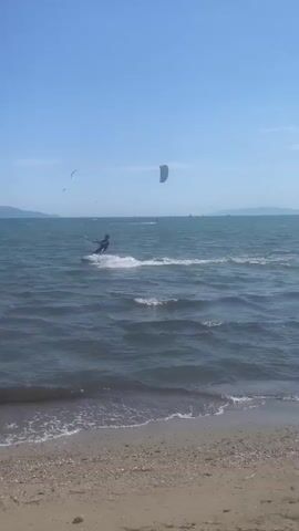 A wide shot of a beach with a person kitesurfing in the ocean under a clear blue sky. The kitesurfer is visible in the center of the frame, leaning back and kicking up spray. In the background, there are other kitesurfers and windsurfers, as well as a distant shoreline with hills. The waves are small and gentle, lapping at the sandy beach in the foreground. The sun is shining brightly, casting a warm glow on the scene.