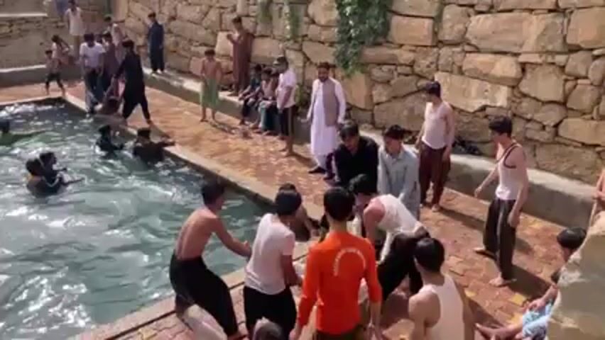 The video shows a group of young men and boys around and in a small, stone-lined pool. Several people are in the water, some swimming, while others are on the side. Several people push a person in black clothing into the pool. Some are dressed in traditional clothing, while others wear casual attire. A person in an orange shirt with lettering is present. The environment includes a stone wall as a backdrop. The atmosphere appears playful and lighthearted.