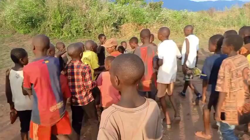 The video shows a group of about 20 African boys gathered outside in a grassy area. Some of the boys are wearing soccer jerseys. They appear to be listening to instructions from an older male standing to the right of the frame. Mountains can be seen in the distance.