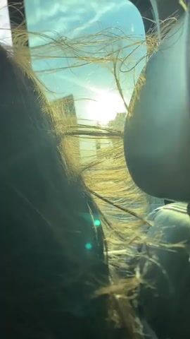The video shows a close-up view from inside a moving vehicle. A person with long brown hair is sitting in the back seat, and their hair is blowing rapidly, obscuring the view out the window. Through the window, a bright sun is visible along with parts of buildings and a blue sky with some clouds. A gray headrest is partially visible on the right side of the frame. The overall impression is that the vehicle is traveling at a high speed with the window open.