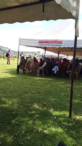 The video shows a group of people sitting in chairs under a tent in a grassy outdoor area, possibly attending an event. There are two tents visible, one larger and one smaller. The larger tent has the words "JNP TENTS, CHAIRS & PA SYSTEM" printed on it. Some people are seated under the smaller tent closer to the camera. The overall scene suggests an outdoor gathering or ceremony.