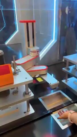 The video shows a close-up view of a robotic arm system enclosed in a clear display case. The arm is silver with red accents and moves along a track. Nearby is a platform with a yellow and red item on it. A person is shown placing their hand in a square opening in the display case, seemingly interacting with a screen or sensor inside. The background is decorated with silver and blue geometric shapes and neon lighting. The scene appears to be part of an interactive exhibit or demonstration showcasing automation or robotic technology.