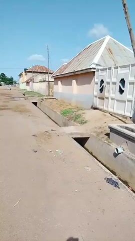 The video shows a slow pan of a dirt road in a village under a clear blue sky. On the right side of the road is a building with a light pink facade and a corrugated metal roof. The building has a large, ornate white metal gate. A concrete drainage ditch runs alongside the road in front of the building. Further down the road, other buildings with corrugated metal roofs are visible. The road is uneven and has several puddles. In the distance, there are more buildings and a few sparse trees. The camera moves slowly from right to left, showing more of the road and the surrounding environment. At one point, a figure can be seen walking in the distance on the left side of the road.