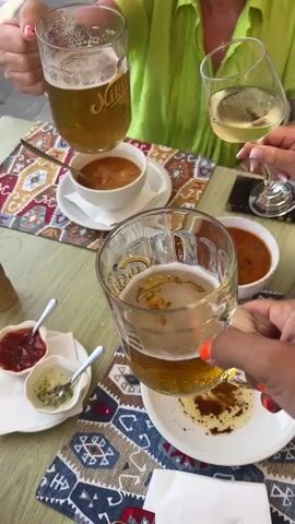 A group of women are enjoying drinks at an outdoor cafe. Two women are holding up beer mugs and one is holding up a wine glass in a toast. The table is set with plates of food and condiments. In the background, a street scene is visible with a car, potted plants, and people walking by. The cafe appears to be located near a body of water, possibly a lake or the sea, with trees and hills in the distance.