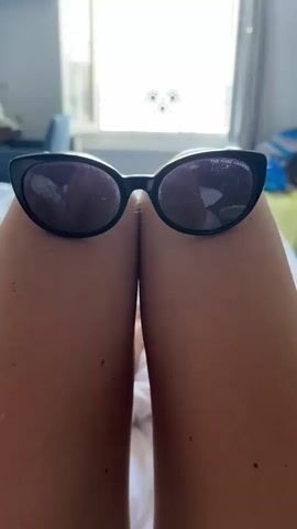 The video begins with a close-up of a pair of black cat-eye sunglasses resting on someone's bare thighs. The camera then pulls back to reveal a bedroom setting with a large window open to a bright, sunny day. Through the window, a view of the sea and a Ferris wheel can be seen. The camera continues to zoom in on the window, showing more detail of the beach, the railing in front of the window, and the ocean. The video ends with a zoomed-in view of the railing and the ocean.