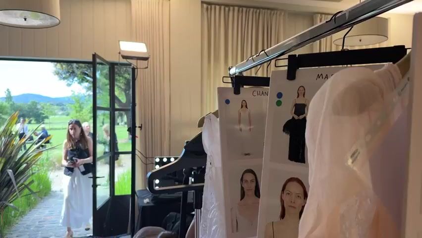 A room appears to be backstage at a fashion show, filled with clothing racks and preparation materials. A rack displays images of models in various outfits. One image shows a woman named Kiko wearing a light pink, floor-length dress with ruffles. A corresponding dress is hanging nearby. Several people mill around the room, including a woman in a white skirt entering from outside. A man with a beard is talking with another woman. There are lighting rigs and other equipment, indicating a professional setting. Outside, a glimpse of greenery and other people can be seen through an open doorway.