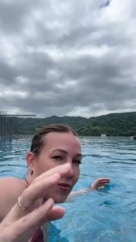 A woman with her hair in a bun swims in an outdoor pool with a cloudy sky and hills in the background.  The camera follows her until it is interrupted by a man who comes into the frame with a surprised expression on his face.