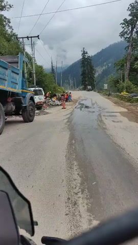 The video shows a road in a mountainous region. There are several people working on the side of the road, wearing orange vests. Construction materials and equipment are visible, including a white vehicle. The road appears wet in places. A dark vehicle, possibly a motorcycle, is in the foreground, suggesting the video is recorded from a moving vehicle. The sky is cloudy, and the surrounding landscape is characterized by steep, tree-covered mountains. Power lines run along the side of the road.
