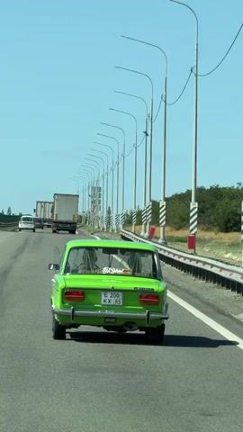 A bright green, vintage sedan drives down a multi-lane highway under a clear blue sky. The car is the central focus, positioned in the right lane of the highway, seen from the rear. It has a license plate with Cyrillic characters and numbers. Several other vehicles, including trucks and cars, are visible further down the highway, traveling in the same direction. The highway is lined with tall, slender streetlights on the right side, each with a black and white striped base and a curved arm holding the light fixture. A metal guardrail runs alongside the streetlights. The landscape beyond the highway features sparse green vegetation and trees. The perspective suggests the video is filmed from another vehicle traveling behind the green sedan. The overall scene depicts a typical highway travel scene.