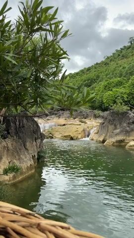 A slow-motion video captures a serene natural landscape. In the foreground, lush green foliage hangs from a branch, partially obscuring the view. The middle ground features a tranquil pool of green water, reflecting the overcast sky. Beyond the pool, a series of gentle waterfalls cascade over beige rock formations. The background is dominated by a verdant, tree-covered mountain range under a cloudy sky. The overall impression is one of natural beauty and peacefulness. There are no people or explicit actions depicted in the video.