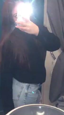 A young woman takes a selfie in a mirror. She is wearing a black top and light-wash jeans. The room appears to be a bathroom. The flash from her phone reflects brightly in the mirror.