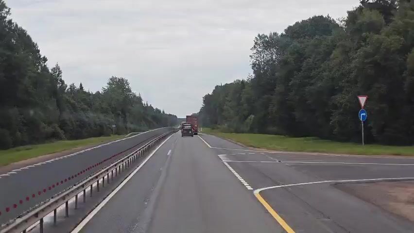 A first-person perspective video shows a truck driving on a highway. The truck is red and appears to be carrying cargo. The highway is surrounded by trees on both sides. The sky is cloudy. The truck is in the right lane and there are no other vehicles visible in the immediate vicinity. The truck is moving at a moderate speed.