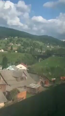 The video shows a green landscape with houses and hills under a cloudy sky. A train can be seen crossing a bridge in the distance. A few people are visible in a grassy area. The video seems to be taken from an elevated position, likely from inside a moving train.