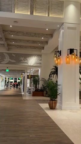 The video pans across a hotel lobby. In the foreground, a wooden floor leads to a large white pillar adorned with wall sconces holding amber glass shades. Potted plants are placed at the base of the pillar. To the left, a hallway with people walking and a dining area with tables and chairs are visible. The ceiling features exposed beams and white louvered panels. In the background, white gazebos with palm tree decals and hanging swings are set up in a lounge area. People are seated in the lounge area. The overall atmosphere appears to be a tropical-themed resort lobby.