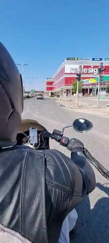 The camera angle shows a person riding a motorcycle in a street during the day. The person is wearing a helmet and a black leather jacket, and is holding the handlebars. A cell phone is affixed to the handlebars for navigation purposes. The street appears empty except for one car far ahead. Tall buildings are visible on the right side of the street, some with the word "redesign" displayed prominently. The sky is clear and blue. The overall impression is one of a sunny day with light traffic on the road.