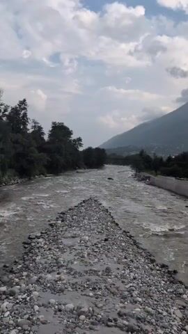 A video begins with a wide shot of a fast-flowing river in a mountainous region. The river is rocky, with a clear path of stones leading into the water in the foreground. Trees line the left bank, and a mountain looms on the right. The sky is partly cloudy. The camera then pans to the left, showing a closer view of the trees and the rough river surface. It then shifts to follow a young man with a backpack walking away from the camera on a paved road. The road is bordered by a concrete barrier with black and white stripes. The man is walking towards a bridge, and behind him, the landscape consists of lush green hills dotted with houses. The video ends with a view of the bridge and the surrounding hills.