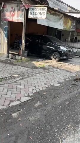 The media file shows an exterior scene of a car service shop, likely a mechanic or car wash. A black car is parked inside an open garage space. Two individuals are visible, one appearing to be working on the car and the other standing nearby. The shop has a sign above the entrance that reads "SERVICE DINAMO BAGONG" and below it, "JL. Raya Karangrejo No. 5". The shop front has various posters and advertisements. The ground in front of the shop is paved with interlocking bricks and is somewhat wet and dirty. The scene appears to be taking place during the day, likely under sunny conditions given the shadows. The overall setting is a commercial street in Gelam, East Java, characterized by modest shopfronts and utilitarian services. The activity suggests routine maintenance or repair work on the vehicle.