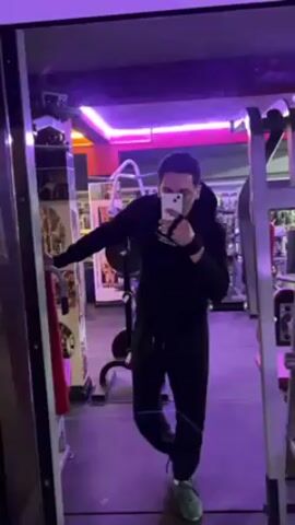 In a gym with purple neon lighting, a person stands in front of a mirror, taking a selfie with a white smartphone. The person is wearing a black hoodie, black sweatpants, and green sneakers. They also have glasses and a black wristband. The person leans against the mirror frame with their left arm and holds the phone with their right hand. Behind the person, there are various gym equipment and another individual in the background.