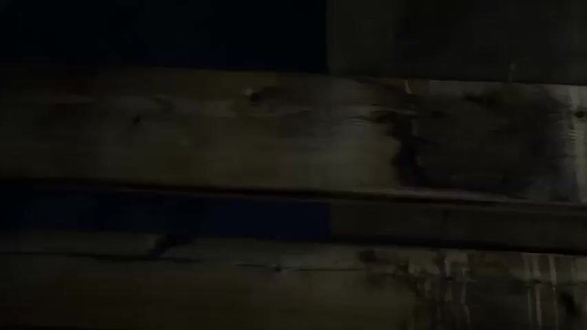 The video shows a dark, grainy, and somewhat blurry view of the night sky seen through a gap in wooden planks. The moon is visible as a bright, circular light in the center of the frame, partially obscured by dark, wispy clouds. The wooden planks are rough and appear weathered. The overall impression is of looking through a window or opening at a nighttime scene.