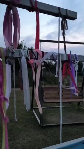 A wooden swing set is in the foreground with several colorful fabric streamers hanging from the crossbeam. The streamers are of various colors and lengths, including pink, red, white, purple, and yellow. Some of the streamers are frayed or torn. In the background, there is a wooden bench swing. The setting appears to be outdoors with grass and trees visible. The sky is overcast.