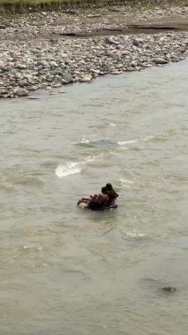 Two individuals are seated on a black inner tube in a wide, murky river. They are positioned facing away from the camera, with one individual behind the other. The river has visible currents and ripples. The riverbanks are lined with rocks and gravel on the left side, with a gentle slope leading to vegetation and buildings in the distance. On the right side, a brick structure is partially visible in the foreground, and leafy branches obscure some of the background. The overall scene depicts a recreational activity in a natural river environment.