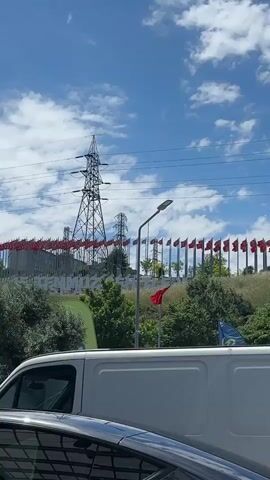The video shows a view from a moving vehicle, possibly a car, of a roadside monument in Turkey under a cloudy sky. The monument is for "15 TEMMUZ SEHITLERI" (July 15th Martyrs), surrounded by Turkish flags. A large electrical transmission tower is behind the monument, and trees and shrubs are in the foreground. A white van is partially visible on the left side of the frame, closer to the camera.