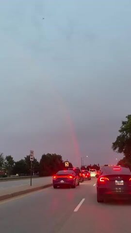 A vertical shot captures a road with heavy traffic under a cloudy sky. A faint, red-hued rainbow arches across the sky, its top visible near a small airplane. Cars are stopped in lanes, their brake lights illuminated. A one-way sign is visible on the left side of the road. Trees line the side of the road.
