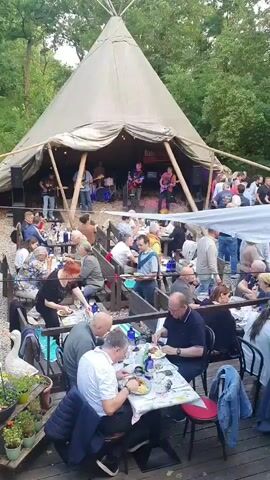 A live band performs on a stage inside a large, conical tent. Numerous people are seated at tables, eating and drinking, arranged in rows in front of the tent. The setting appears to be outdoors, with trees and foliage visible in the background.