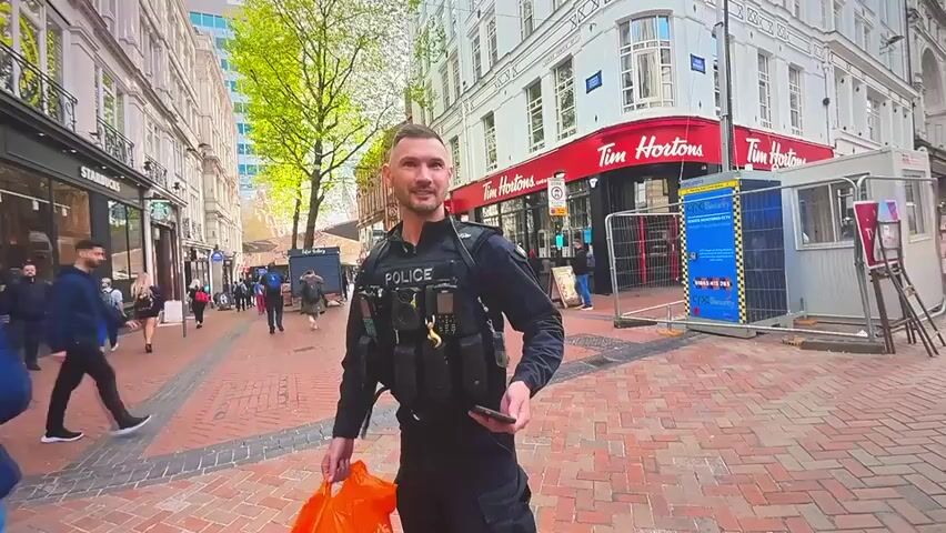 The video shows a street scene with a police officer in the foreground. The officer is wearing a dark uniform with a vest that says "POLICE" on it. He is holding a phone and an orange shopping bag. In the background, there are buildings, shops, and other people walking on the street. One building has a large red sign that says "Tim Hortons". There is also a police car visible in the last few seconds of the video. The car is white with blue and yellow police markings.