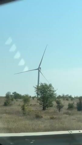 A person is filming a video from a moving vehicle, possibly a car, as indicated by the partial view of a vehicle's interior at the top of the frame and the blurry nature of the landscape in the distance. The video shows a series of wind turbines dotting a dry, grassy landscape with scattered bushes and small trees. The sky is a clear, pale blue with a few wispy clouds. The wind turbines are tall, with white blades, and appear to be functional. The vehicle appears to be traveling on a road with a guardrail visible in the foreground. The overall scene depicts a wind farm in a rural, possibly semi-arid environment.