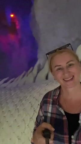 A blonde woman in a plaid shirt and sunglasses on her head smiles as she films herself with a selfie stick. She is in front of a white, dragon-like sculpture with intricate details. The environment appears to be dimly lit with purple and red lighting in the background. The floor is a light color, possibly sand or gravel.