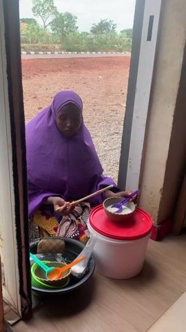 A young woman wearing a purple hijab sits on the floor preparing food. She is next to a bucket with bowls and spoons, and a white bucket with a red lid. She is in a doorway and the outside area appears to be gravel. She is using a stick and a purple spoon to prepare the food in the bowl.