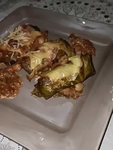 A plate of stuffed cabbage rolls is shown in the video. They appear to be covered in melted cheese and sauce. One roll is also topped with shredded cheese.  The plate is rectangular and light gray in color. The background features a tablecloth with a black and white pattern.