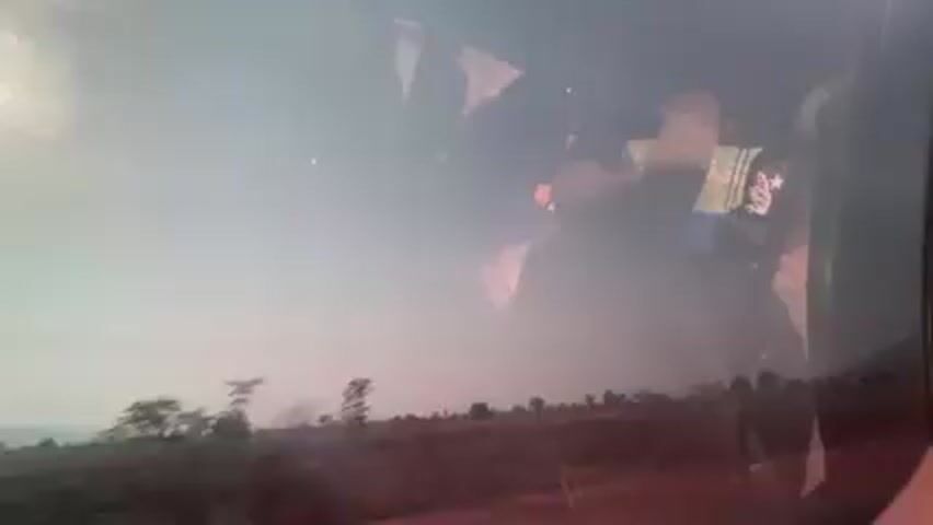 The view from inside a moving vehicle shows a rural landscape through the window. The landscape appears dry and dusty, with sparse vegetation and telephone poles lining the roadside. There are people walking along the side of the road. The sky is hazy and the overall lighting suggests either early morning or late afternoon. The reflection in the window partially obscures the outside view, but it shows the interior of the vehicle and the people within. The camera seems to be held by someone inside the vehicle, capturing the passing scene.