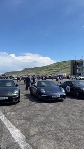 The video shows several parked Porsches at an outdoor event with a crowd of people in the background. The sky is clear and blue, and there is a hill in the distance. The camera pans slightly to the right, revealing more cars and people. The Porsches are various colors, including gray, blue, and green. One of the cars is marked with the number "77" on its side. There is a building in the background, possibly related to the event. The atmosphere appears casual and sunny.