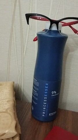 A pair of black-rimmed glasses with red arms are balanced on top of a tall, dark blue bottle. The bottle has a white cap and a label that reads "PRINCESSESSEX" and "9% OXYGIANT" in white text. The bottle is standing on a wooden surface, likely a table or shelf. To the left of the bottle, a beige, textured fabric object is partially visible. The background is a pale, textured wall with vertical lines. The lighting appears to be artificial and casts soft shadows. The scene is static, with no visible movement.