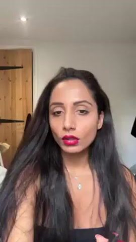 A woman with long, dark hair is applying dark red lipstick. She is fair-skinned and is wearing a nose ring and two necklaces. The background includes a white wall, a light-colored wooden door, and a black garment hanging on the wall behind her. A light fixture is visible in the upper left of the frame.