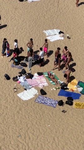 The video shows a bird's-eye view of a sandy beach scene with a group of teenagers and their belongings spread out. There are approximately 14 individuals, mostly young adults, standing and milling around a collection of towels, bags, clothing, and personal items. Some are in swimsuits or shirtless, suggesting they are prepared to swim or sunbathe. One person is lying on a green towel, seemingly sunbathing.  There are several colorful beach towels with various patterns spread on the sand. Backpacks and other bags are scattered around. A few other individuals are visible in the periphery. The weather appears sunny and the scene is typical of a casual beach gathering.
